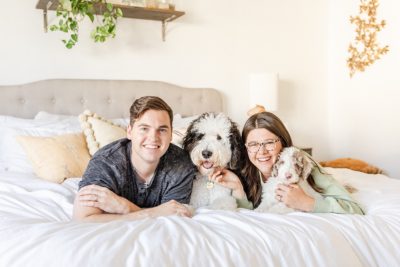 parents sit on bed with Sheepadoodle dog and Sheepadoodle puppy