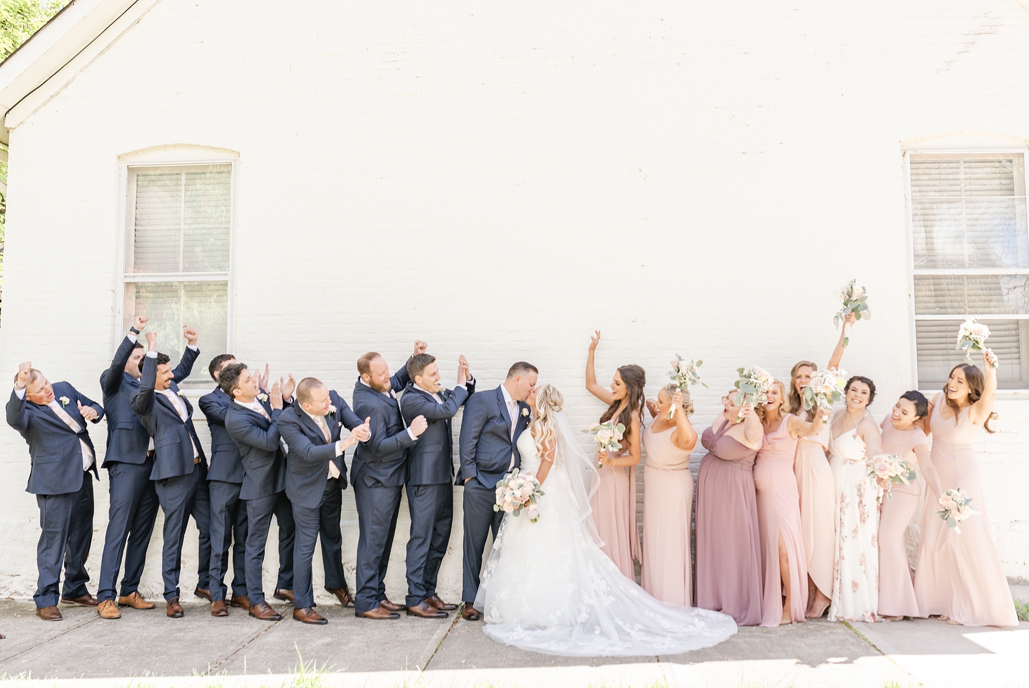 bride and groom kiss by white wall with bridal party cheering